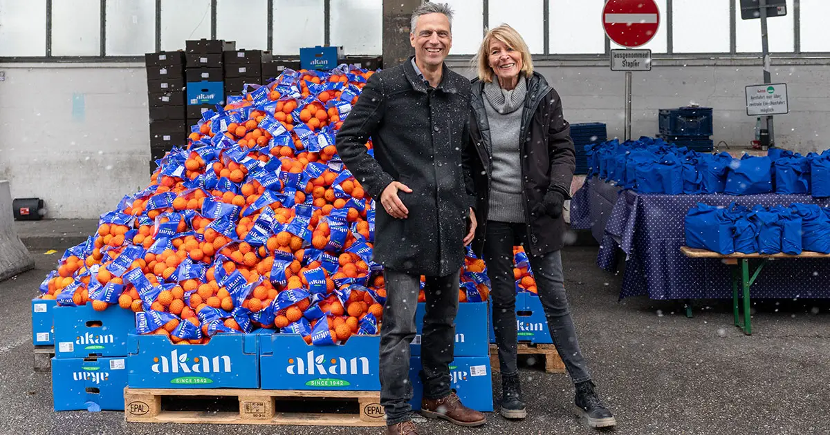 Rocco Bräuniger, Country Manager Amazon Deutschland und Hannelore Kiethe, Vorstandsvorsitzende der Münchner Tafel. Beide stehen vor der Ausgabestelle am Großmarkt, vor vollen Orangenkisten.